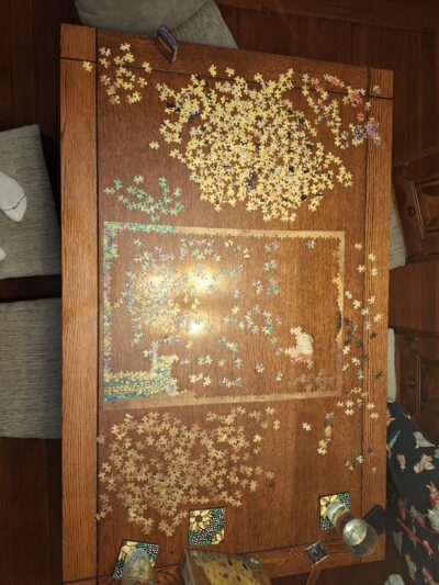 A partially completed jigsaw puzzle of Klimt’s “The Kiss” laying on a wooden dining table, with the edges and the bottom right corner of flowers finished.
