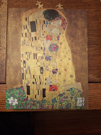 A jigsaw puzzle box showing the puzzle (Klimt’s painting “The Kiss”) laying on a brown dining table.