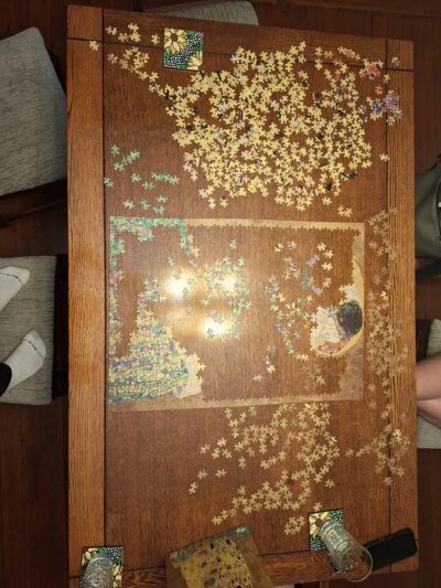 A partially completed jigsaw puzzle of Klimt’s “The Kiss” laying on a wooden dining table, with the lover’s faces and the bottom right corner of flowers finished.
