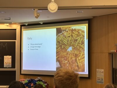 A picture showing a slideshow on a projector screen. The slide is titled Italy and describes "Pizza Americana" with cheap hot dogs and french fries. There is a picture of the "Pizza Americana" to the side.