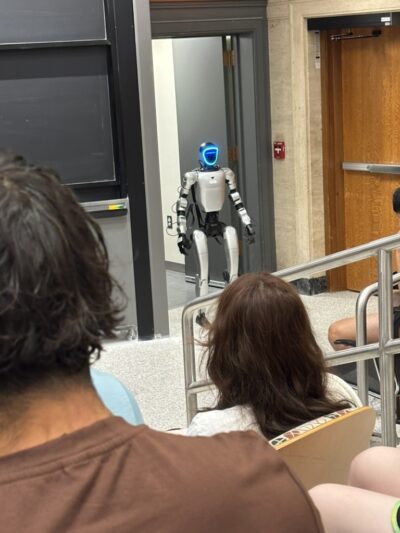 A picture taken from inside a lecture hall. There is a gray humanoid robot standing by the door.
