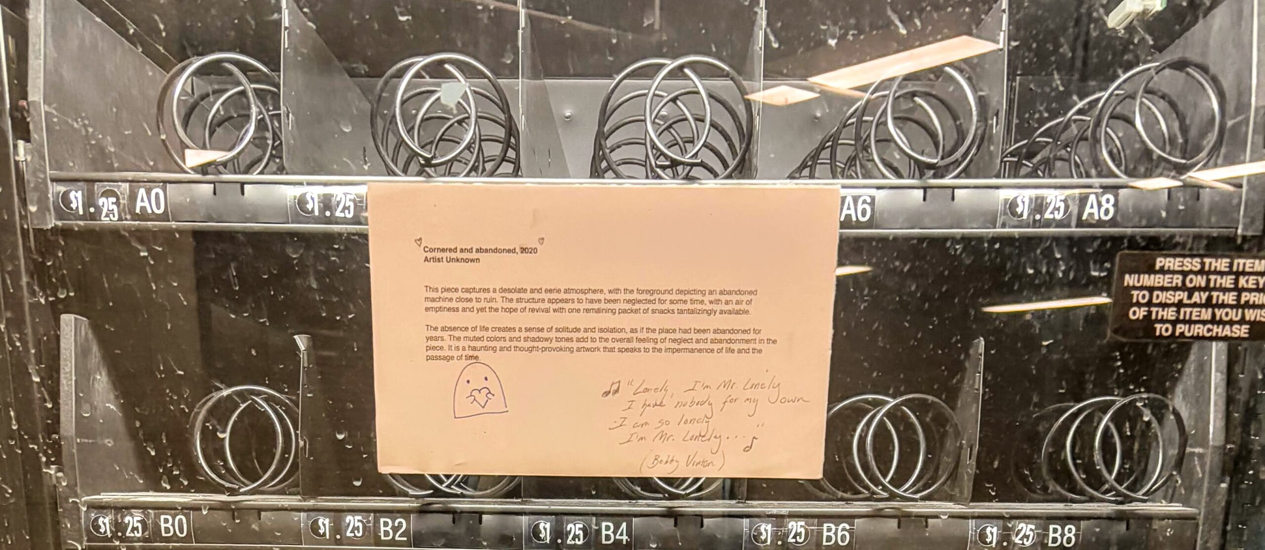 top rows of a vending machine with a placard pasted on the outside in parody of an art museum sign titled "cornered and abandoned, 2020 / artist unknown" that describes the desolate abandonment of the vending machine