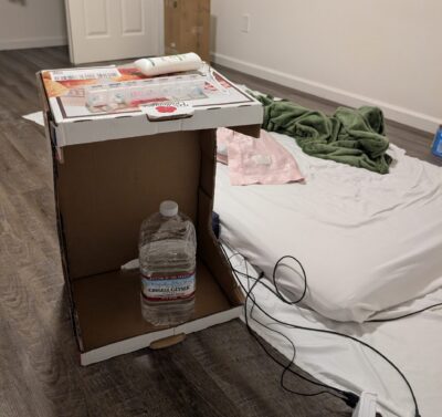 open-mouthed cardboard box stood on its side, with assorted items on the top surface and a large waterbottle on the bottom surface, with a futon mattress in the background