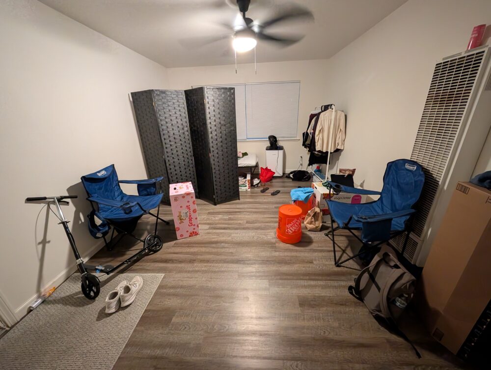 large room with two camping chairs, upside down home depot buckets, cardboard boxes, a clothes rack, a room divider, and a bed behind the divider. quite messy