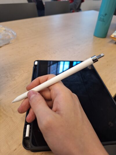 image of a white apple pencil with the end cap off, exposing the charging port. a cased iPad lays below on a table with the screen exposed but off.